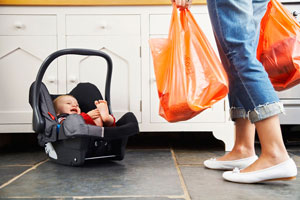 Woman carrying grocery bags with a baby sitting in a carrier on the ground by the woman's feet.